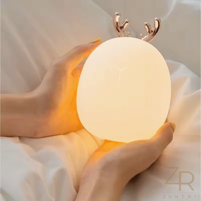 Happy girl lying in bed, smiling at a glowing bunny night light that creates a comforting bedtime atmosphere.